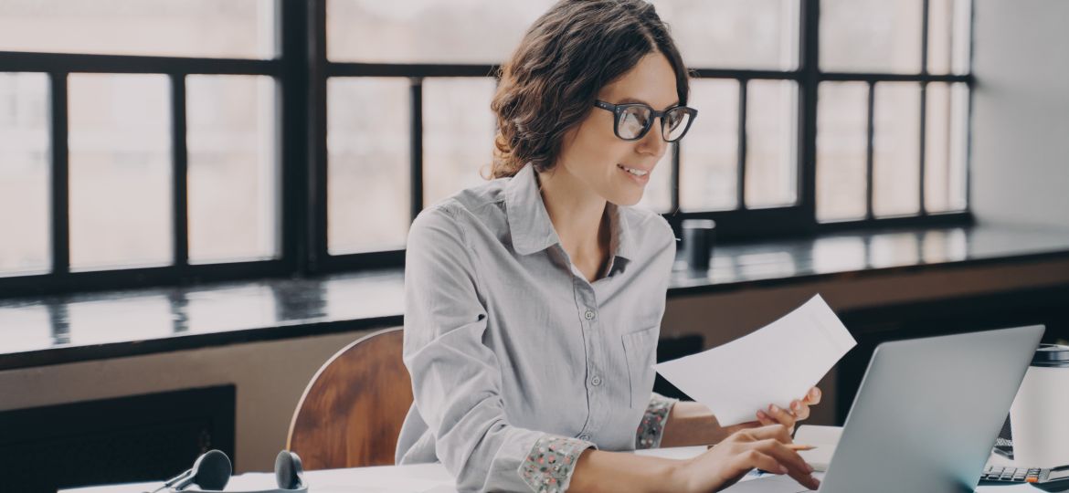 Focused European business woman sitting at home office holds financial documents, working on laptop