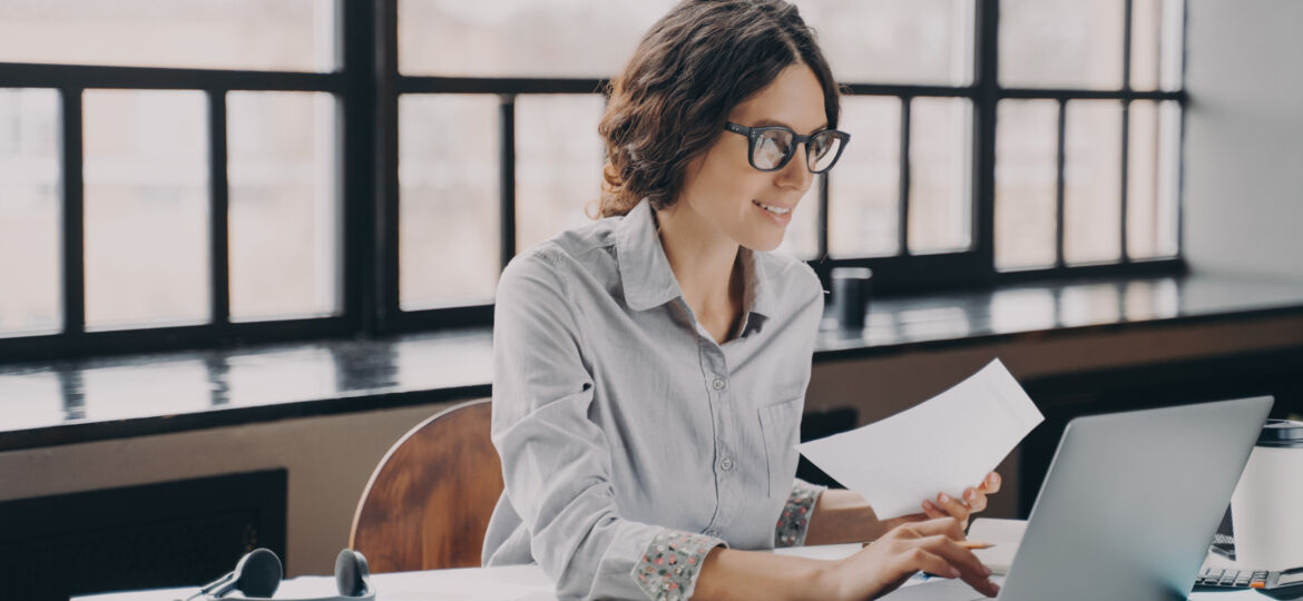 Focused European business woman sitting at home office holds financial documents, working on laptop