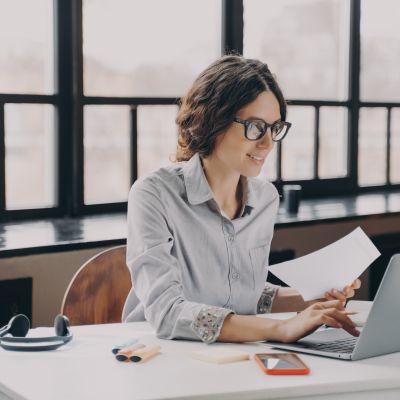 Focused European business woman sitting at home office holds financial documents, working on laptop